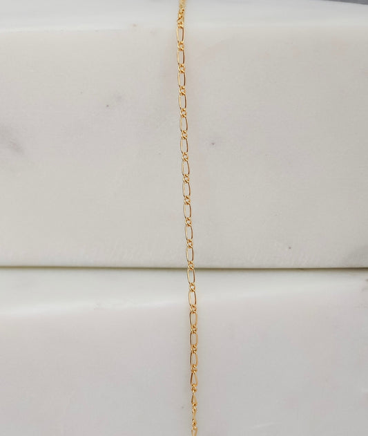 "Delicate gold chain displayed vertically on a white marble block, showing its small oval links."