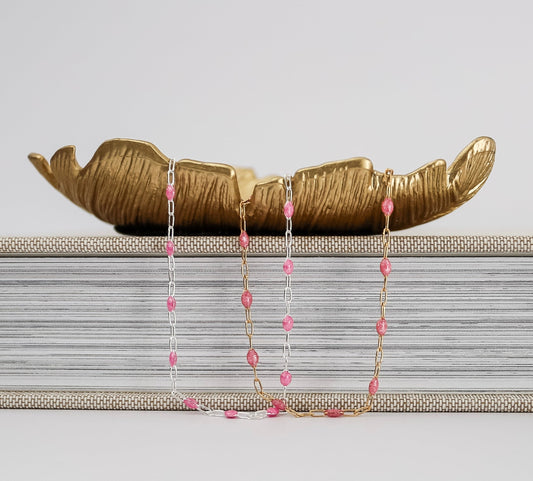 Delicate gold and silver chain necklaces with small pink gemstone beads, draped over stacked books with a decorative gold leaf accent on top.