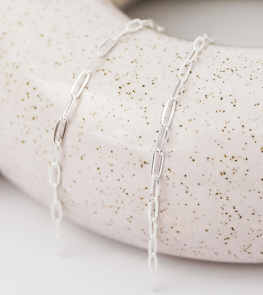 Close-up of a delicate silver chain featuring small open diamond-shaped links, displayed draped over a light speckled ceramic surface.