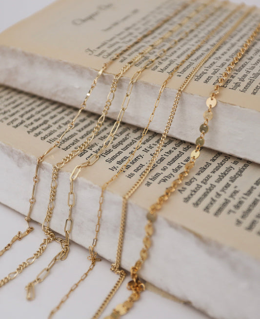 Here’s an SEO-friendly and descriptive alt text for the image:  "Various styles of gold-filled chains laid across open book pages, showcasing different link patterns and textures for permanent jewelry making